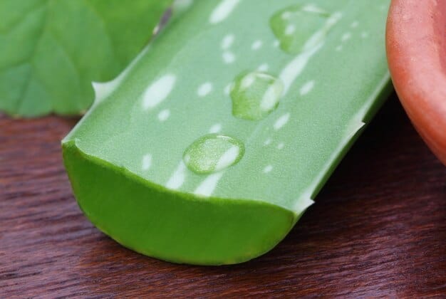 A freshly cut Aloe Vera leaf with visible inner gel and water droplets on its surface, placed on a wooden table.